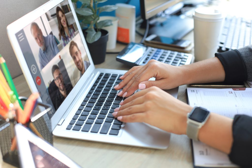 Person tippt auf einem Laptop, auf dessen Bildschirm eine Videokonferenz mit vier Teilnehmenden zu sehen ist; auf dem Schreibtisch stehen unter anderem ein Smartphone, ein Notizbuch, ein Kaffeebecher und eine Pflanze.