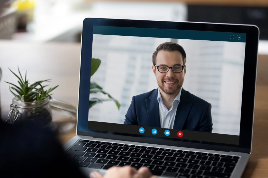Laptop mit geöffnetem Videoanruf: Eine Person in einem Anzugjackett ist auf dem Bildschirm zu sehen; am unteren Rand sind Symbole für Video‑, Audio‑ und Anrufsteuerung sichtbar, im Vordergrund liegt eine Hand auf der Tastatur.