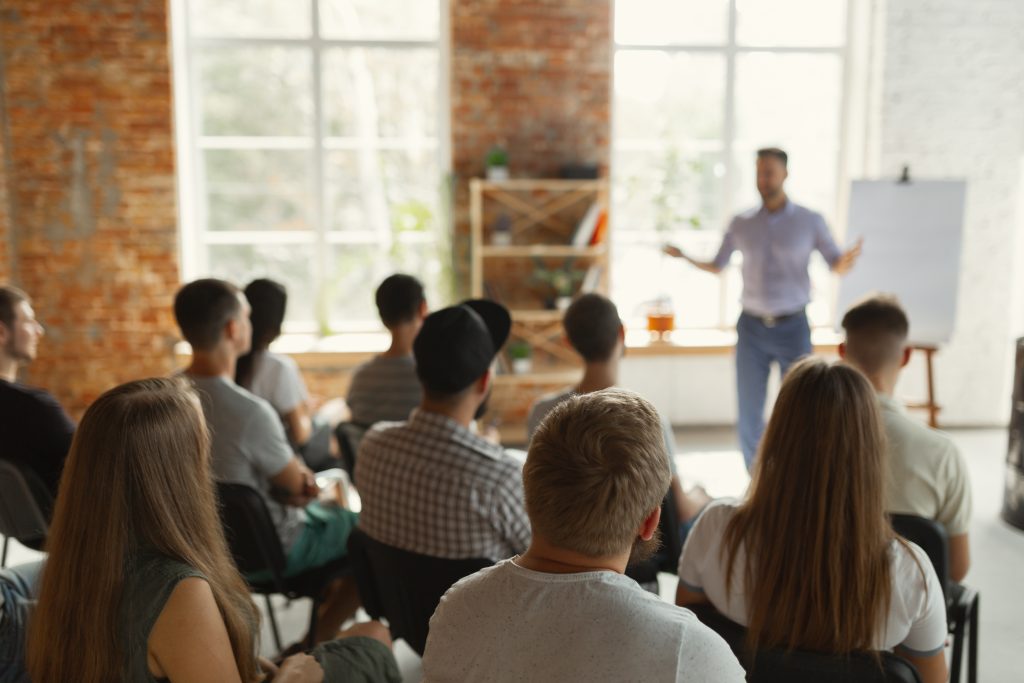 Mehrere Personen sitzen auf Stühlen in einem hellen Raum mit großen Fenstern und Backsteinwänden und schauen nach vorne, während eine stehende Person im Vordergrund eine Präsentation vor einer Flipchart hält.
