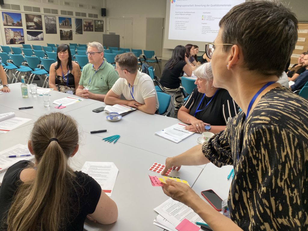 Teilnehmende eines Workshops der TANDEM Sachsen Projekte diskutieren gemeinsam an einem Tisch im smac – Staatlichen Museum für Archäologie in Chemnitz über Qualitätsmerkmale und die Weiterentwicklung guter Praxis. Im Hintergrund ist eine Präsentation zu sehen