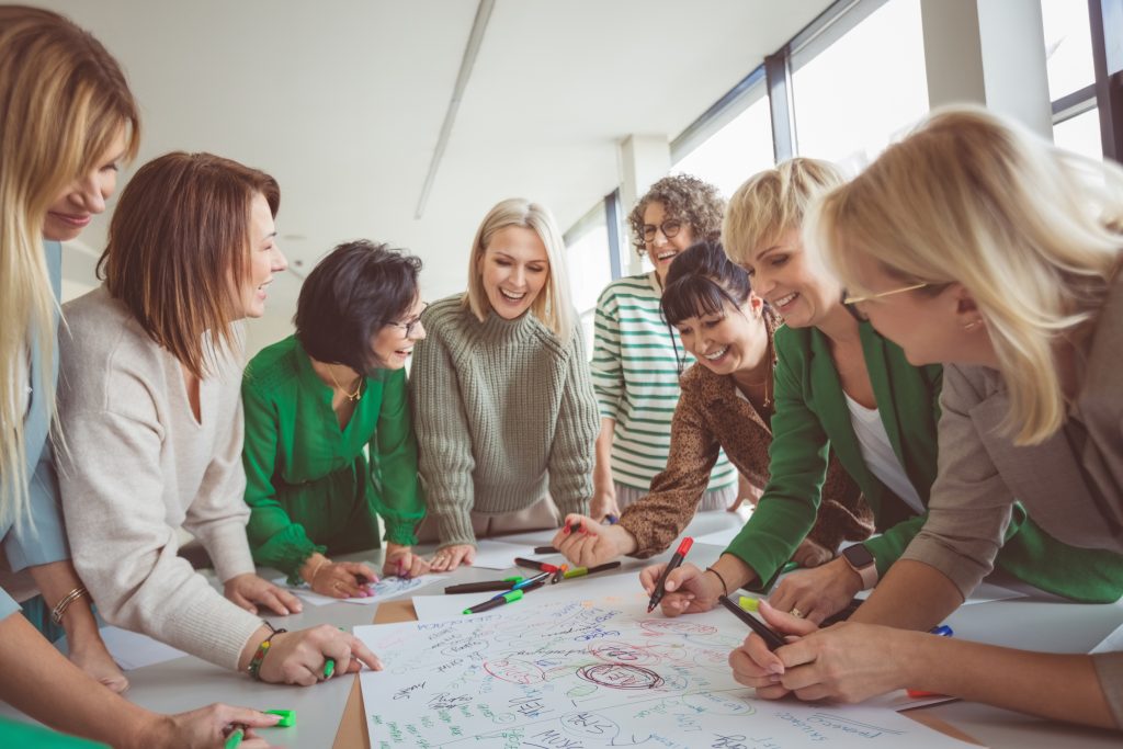 Eine Gruppe von acht Personen versammelt sich um einen Tisch in einem hellen Seminar- oder Besprechungsraum. Auf einem großen Papierbogen werden mit verschiedenen farbigen Markern Ideen, Begriffe und Skizzen festgehalten. Die Teilnehmenden arbeiten gemeinsam an den Inhalten, beugen sich über das Plakat und tauschen sich aus. Die Szene vermittelt einen kollaborativen Arbeitsprozess, beispielsweise im Rahmen eines Workshops, einer Vernetzungsveranstaltung oder einer Gruppenarbeit.